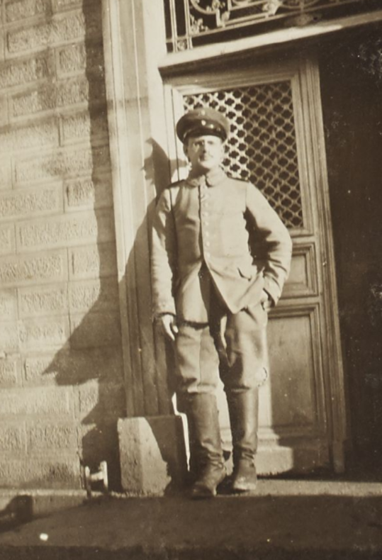 Man sieht Victor Ehrenberg, 1914 als Soldat an der Westfront, wie er in Uniform vor der Tür eines Gebäudes steht